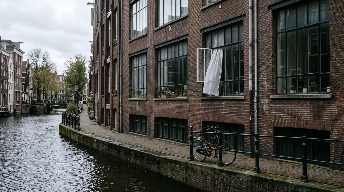 Backsteinhaus am Kanal, große Fenster, ein Fahrrad, graues Licht auf dem Wasser, ein Vorhang bewegt sich