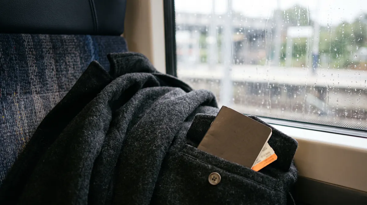 Wollmantel auf einem Zugsitz, Ecke eines geschlossenen Hefts in der Tasche, Regen am Fenster, nichts Lesbares